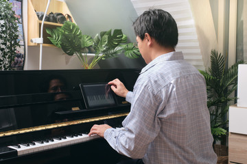 Adult man learning to play piano using digital tablet with an online lesson and course in living room at home, Happy Asian businessman relaxing by playing piano, Reduce stress with music concept