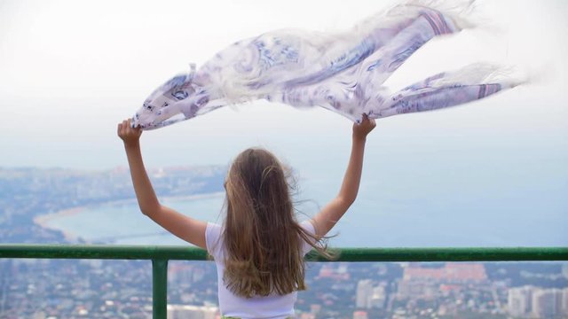 Smiling girl with winding shawl looking at majestic seascape. Back view of cheerful teenage girl standing on observation point and looking at beautiful seascape and coastal town at windy day