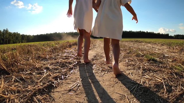 Two little sisters run along the road on the field in the rays of the evening sun in summer in beautiful folk dresses.