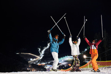 Happy skiers team in winter jackets and helmets raising ski poles and looking at camera, standing...