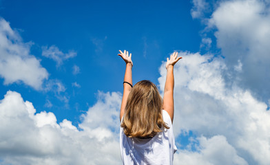 a woman or girl stands with her back to the camera and stretches her hands to the sky. The girl raised her hands up against the blue sky