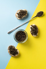 Glass jar and spoon of black caviar and sandwiches with black caviar on blue and yellow background. Top view and copy space