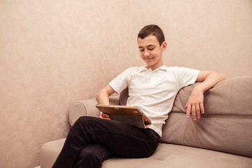 Teenager boy in white T-shirt sits on sofa with laptop
