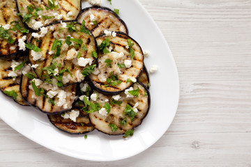 Homemade Grilled Eggplant with Feta and Herbs on a white plate on a white wooden background, top view. Flat lay, overhead, from above. Space for text.