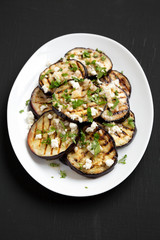 Homemade Grilled Eggplant with Feta and Herbs on a white plate on a black surface, high angle view.