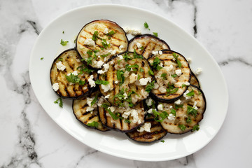 Homemade Grilled Eggplant with Feta and Herbs on a white plate on marble background, top view.