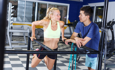 Physically fit female working out triceps and biceps on bars indoors with personal trainer assistance