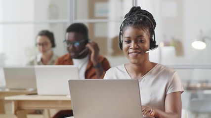 Young African American female agent in headset typing on laptop, smiling and talking on web call while working at desk in call center - Powered by Adobe
