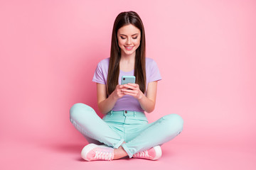 Full length photo of positive cheerful girl sit floor use smartphone repost share subscribe social media news wear turquoise violet purple trousers isolated over pastel color background