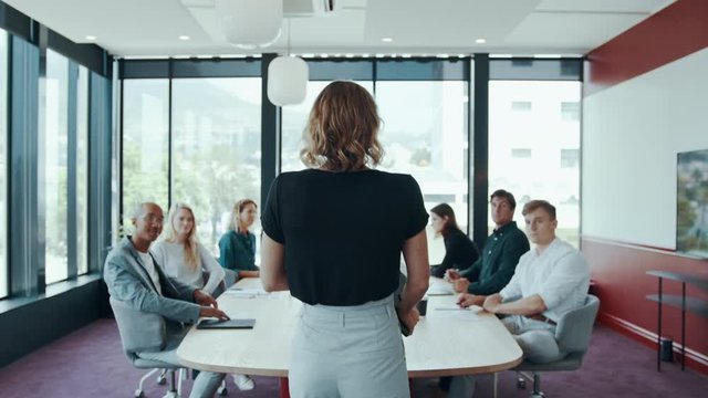 Rear view of a female business leader walking to the conference table with colleagues.  Entrepreneur arriving in the conference room with her team settling down around the table.
