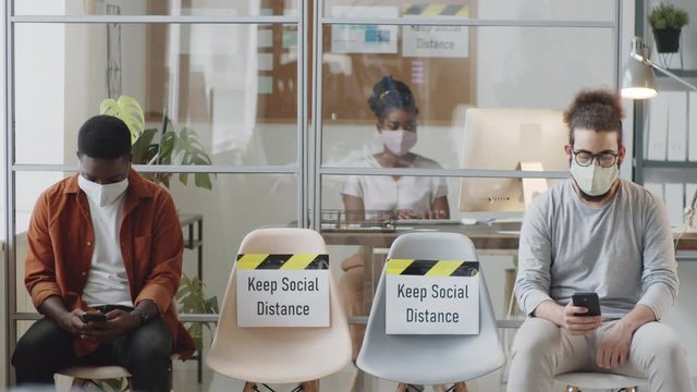 Multiethnic Male Colleagues Sitting Far Apart On Chairs In Office With вЂњKeep Social DistanceвЂќ Posters And Using Smartphones While Afro-american Woman Working On Laptop Behind Glass Wall