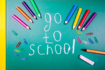 On a green chalkboard it is written to Go to school, next to it are crayons, pencils, colored paper clips, and a pen. Concept of the school, academic year. Beautiful, fun background for school.
