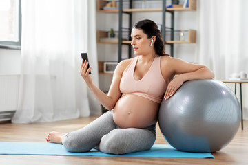 pregnancy, sport and people concept - happy pregnant woman with smartphone, earphones and fitness ball at home