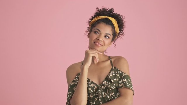 An emotional african american young woman is dreaming about something standing isolated over pink background