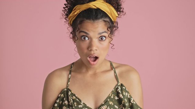 A close-up view of a shocked african american young woman is looking to the camera while opening her mouth standing isolated over pink background