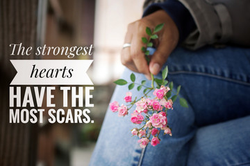 Inspirational words - The strongest hearts have the most scars. Strength motivational quotes concept with young woman hand holding mini pink roses on blue jeans background.