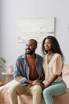young african american husband and wife in casual clothes looking away while sitting on bed