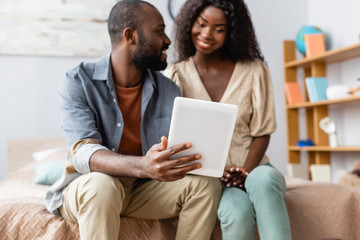 selective focus of african american man showing digital tablet to wife while sitting on bed