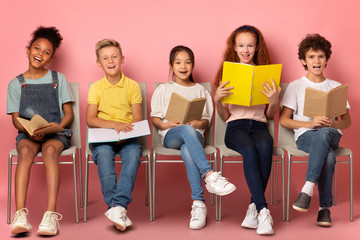 School education. Happy diverse kids with textbooks and notebooks sitting on chairs against pink background
