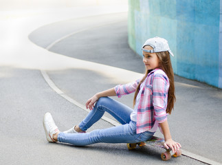 Smiling cute little girl child in cap sitting on a skateboard. Preteen with penny board outdoors