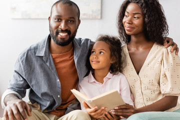 young african american man touching shoulder of wife while sitting near daughter holding book
