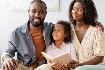 Obraz premium young african american man touching shoulder of wife while sitting near daughter holding book