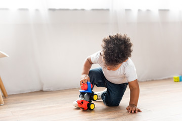 curly african american boy playing with toy truck on wooden floor at home