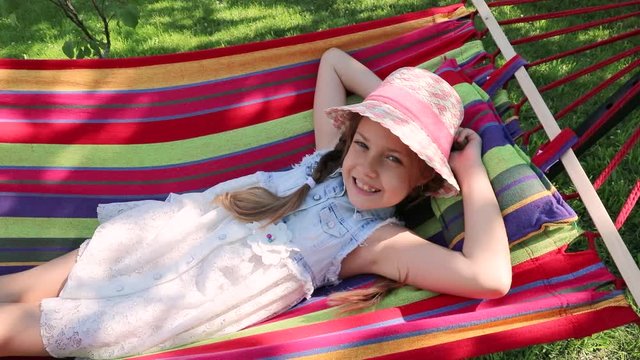 a little girl lying in a hammock on the street in the park. a little sweet girl in a hat lying on a bright hammock. 