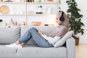 Fototapeta premium Weekend Pastime. Arabic girl relaxing at home with smartphone and wireless headphones