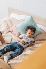 Obraz na płótnie Canvas high angle view of african american boy in white t-shirt and jeans holding tv remote controller while lying in bed