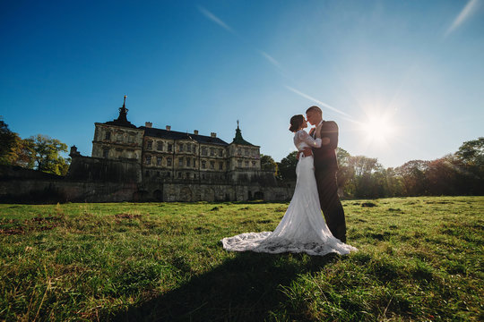 Lovely wedding couple walking in the sunlight near the old castle. Stylish handsome bride and groom in arms against the backdrop of an ancient building and nature
