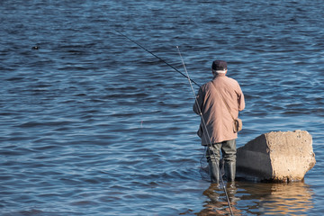 an elderly man in rubber boots is fishing with a line in the water of a river or lake.