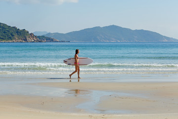 young slender and tanned girl in a swimsuit with a surfboard in her hands on the beach near the water, sea, summer, heat, sunny day, clear sea water, wave, lifestyle, sports, leisure, vacation, weeken