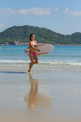 young slender and tanned girl in a swimsuit with a surfboard in her hands on the beach near the water, sea, summer, heat, sunny day, clear sea water, wave, lifestyle, sports, leisure, vacation, weeken