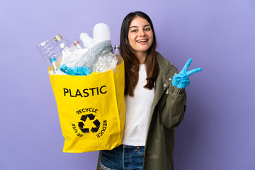 Young woman holding a bag full of plastic bottles to recycle isolated on purple background smiling...