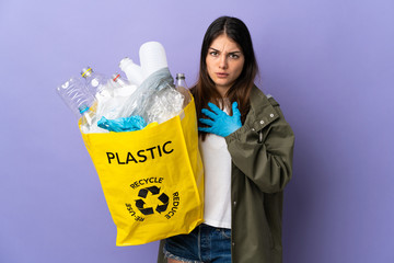 Young woman holding a bag full of plastic bottles to recycle isolated on purple background surprised and shocked while looking right