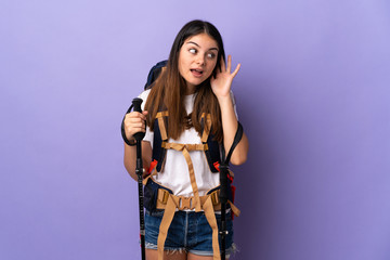 Young woman with backpack and trekking poles isolated on purple background listening to something by putting hand on the ear