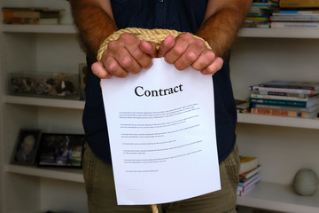 Tied hands holding contract paper in front of bookcase. Bound man hands with paper contract, breach...