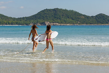 two girls in bekin with a white surfboard enter the blue sea, summer, heat, sunny day, clear sea water, wave