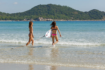 two girls in bekin with a white surfboard enter the blue sea, summer, heat, sunny day, clear sea water, wave