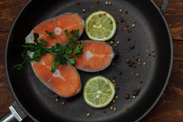 Slices of pink salmon in a black cast iron pan with a bunch of green parsley, lemon and spices top view on a koichnevom wooden background, the concept of cooking lunch, restaurant, healthy food, omega