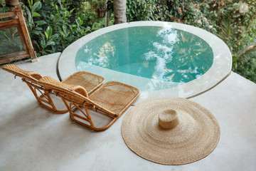 Fototapeta na wymiar Time to chill and relax. Two empty deck chairs near round swimming pool in the jungle, big straw hat lie next to the pool