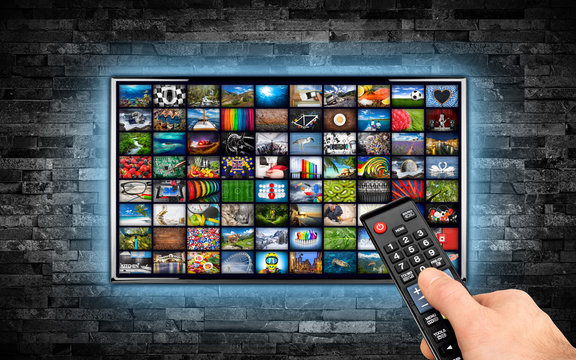 hand with remote on black flat tv screen display with picture movie gallery backdrop. television wall mounted on dark slate stone wall blue LED background. Multimedia streaming internet cloud concept