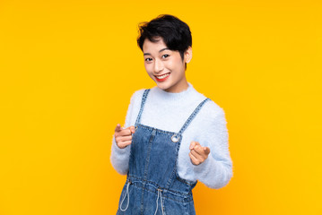 Young Asian girl in overalls over isolated yellow background points finger at you