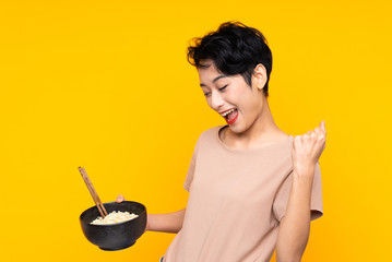 Young Asian girl over isolated yellow background celebrating a victory while holding a bowl of noodles with chopsticks