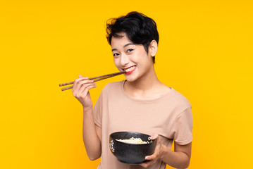 Young Asian girl over isolated yellow background holding a bowl of noodles with chopsticks