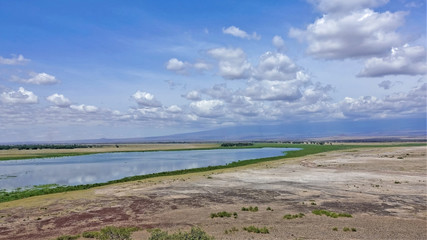 A blue lake on the dry red-brown land of the savannah. Some green vegetation. Sunny day. Fluffy clouds hide the summit of Mount Kilimanjaro and are reflected in the smooth water. Kenya. Amboseli park.
