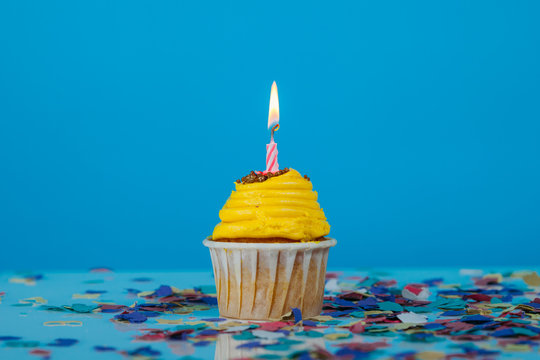 Birthday yellow cupcake. Small muffin or cake with a lit candle. Anniversary celebration with confetti. Horizontal shot, cupcake in the center of the image, on a blue background.