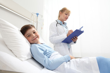 Fototapeta premium Smiling child lying in bed in hospital room looking at camera with doctor beside him filling out his medical record with copy space