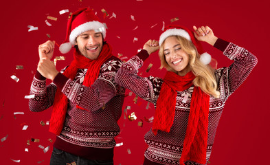 Lovely young couple in sweaters and Santa hats dancing on Christmas or New Year party on red background - Powered by Adobe